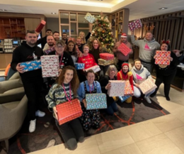 A group photo of people holding presents sat and stood around a Christmas tree 